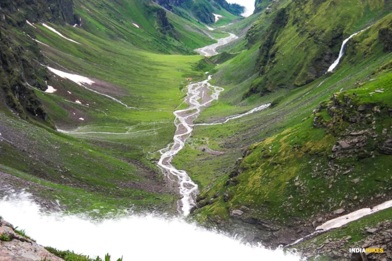 A lush green mountain valley with winding streams and scattered patches of snow enhance the breathtaking landscape, guided by a local Sherpa who unveils the hidden gems of the Himalayas.