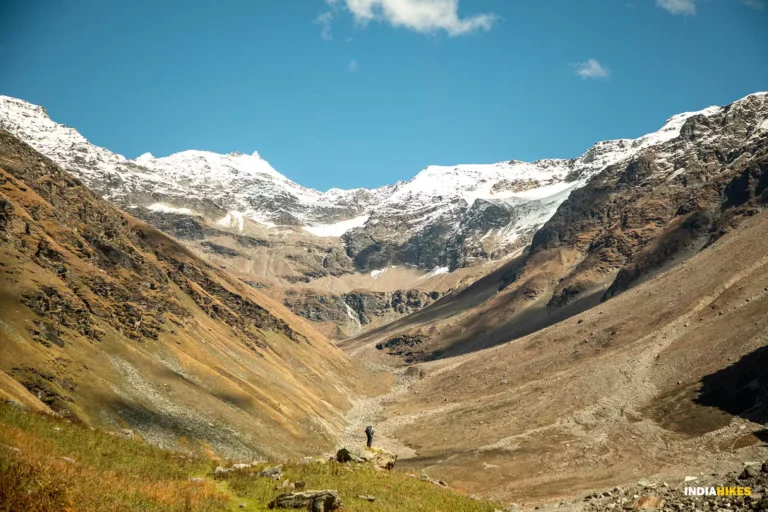 A lone person stands in a vast mountain valley with snow-capped peaks under a clear blue sky, guided by a local Sherpa through the majestic Himalayas.